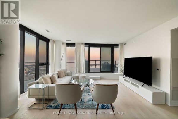 Modern living room with a large TV, white entertainment unit, two grey chairs, and expansive windows showcasing the city. - 4304 - 16 Bonnycastle Street, Toronto