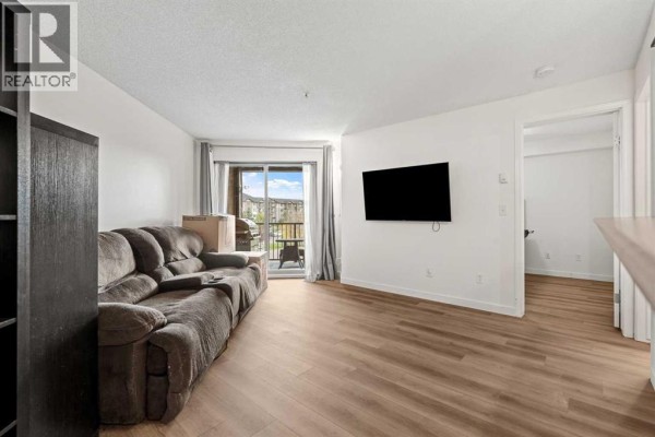 Living room with a brown sofa, wall-mounted TV, and a sliding glass door to a balcony. - 4307, 60 Panatella Street Nw, Calgary