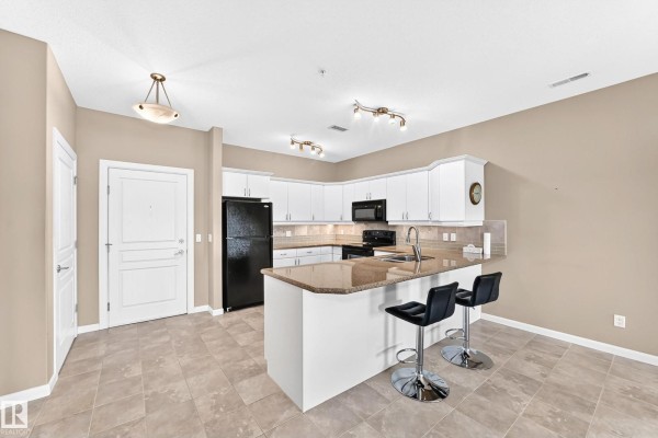 Kitchen and living area with an island, bar stools, white cabinets, black appliances, and a fireplace. - #436 160 Magrath Rd Nw, Edmonton