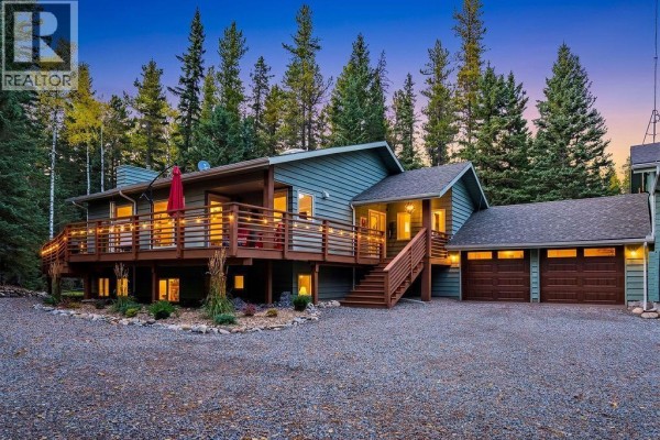 Exterior of the green house and garage lit up at dusk, surrounded by trees. - 44 Elk Valley Grove, Rural Rocky View County