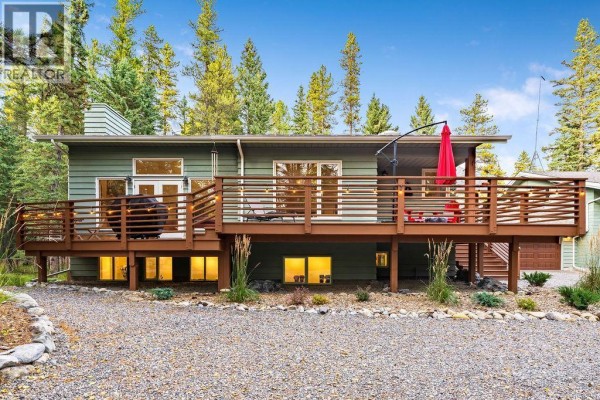 Rear exterior of the green house featuring a spacious wooden deck and views of pine forest. - 44 Elk Valley Grove, Rural Rocky View County