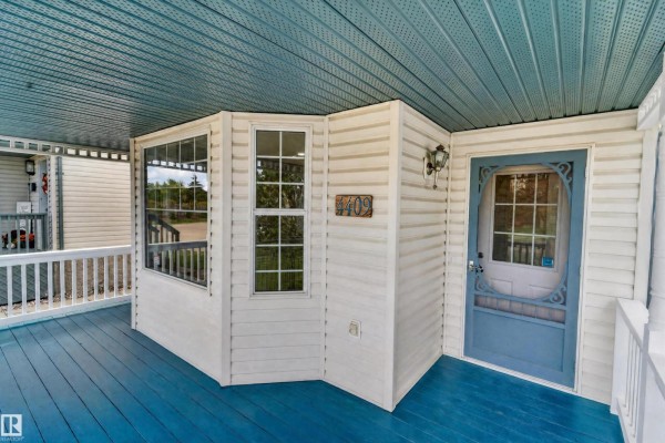 Close-up of the covered porch with a blue door, bay window, and wooden deck. - 4409 150 Av Nw, Edmonton