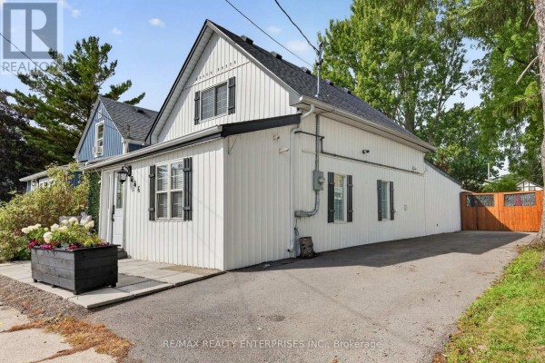 Side and rear exterior view of a white cottage with a driveway and wooden fence. - 446 Cambridge Road W, Fort Erie