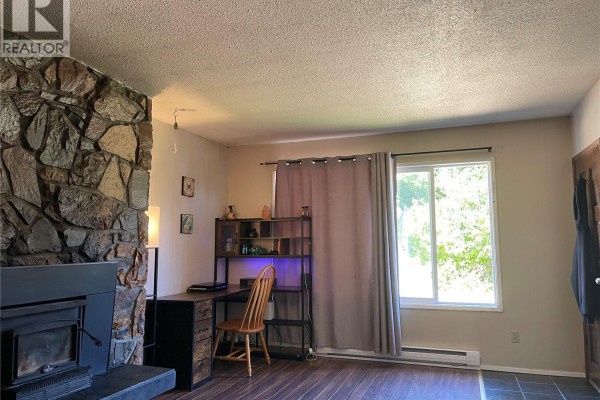 Living room featuring a large stone fireplace, a wooden desk, and a window with grey curtains. - 4475 Byng Rd, Port Hardy