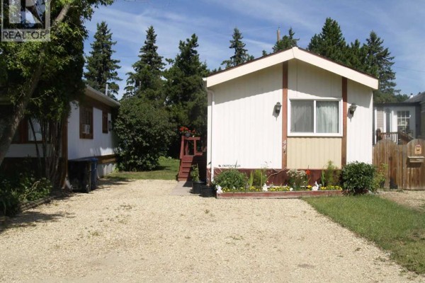 Exterior view of a white mobile home with a red-trimmed entrance and surrounded by gravel and trees. - 44a, 5500 Womacks Road, Blackfalds