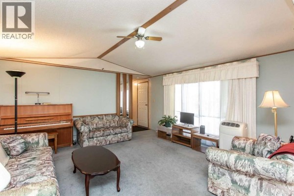 Living room with grey carpet, a piano, two patterned sofas, a coffee table, and a large window. - 44a, 5500 Womacks Road, Blackfalds