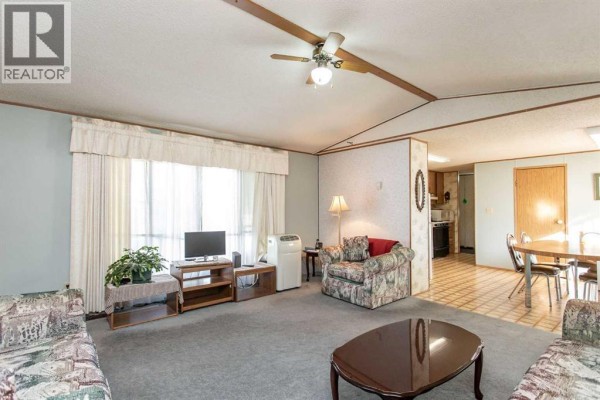 Living room with grey carpet, patterned armchairs, a coffee table, and view into the kitchen/dining area. - 44a, 5500 Womacks Road, Blackfalds