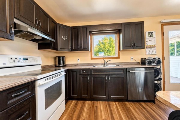 Kitchen counter with dark wood cabinets, stainless steel sink, white oven, and window view. - 45 Bredin Avenue, Rosenfeld