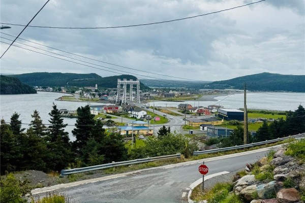 A paved road curves downwards showing a town, bridge, and bay with a stop sign and rocky terrain. - 45 Fort Louis Road, Jerseyside