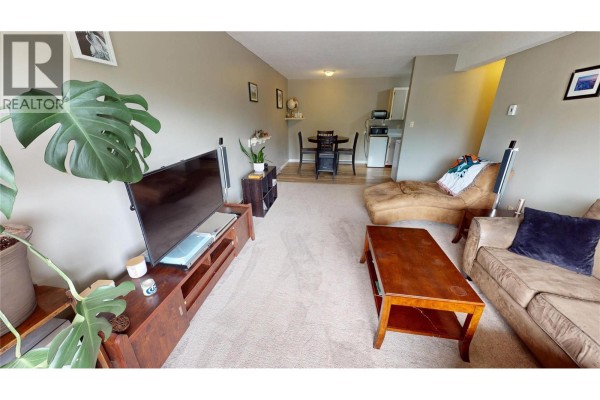 Living room with beige carpet, a large television, and a dining area in the background. - 45 Ridgemont Drive Unit# 302, Fernie