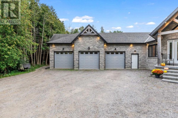 Side view of a stone house with three gray garage doors and a single door, set against a backdrop of trees. - 4500 Old Scugog Road, Clarington