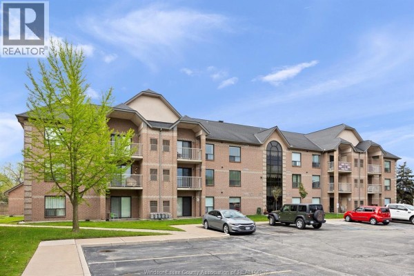 Exterior view of a brick apartment building with balconies and parked cars. - 451 Grand Marais Road West Unit# 114, Windsor