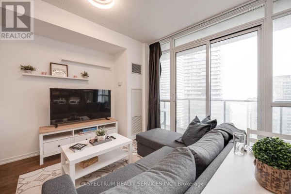 Living room with grey sectional sofa, white coffee table, television, and dining table next to sliding glass doors. - 4511 - 65 Bremner Boulevard, Toronto