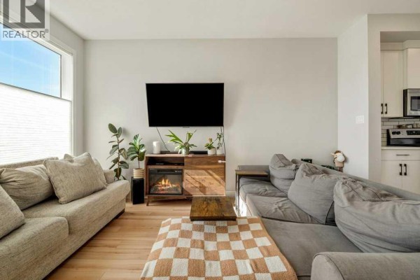 Living room with light brown and grey sofas, electric fireplace, and a view into the open-concept kitchen. - 4531 Fairmont Gate S, Lethbridge