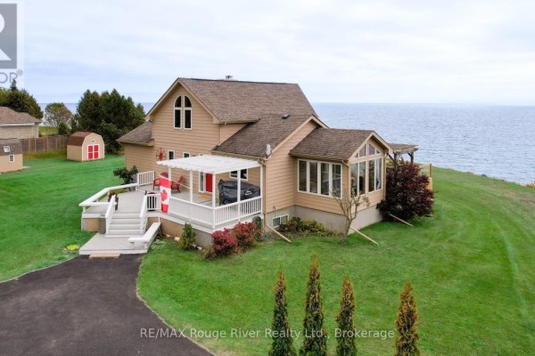 Tan house with a white deck, pergolas, and sunroom, set on a grassy slope beside the water. - 455 Lakeshore Road, Alnwick/haldimand