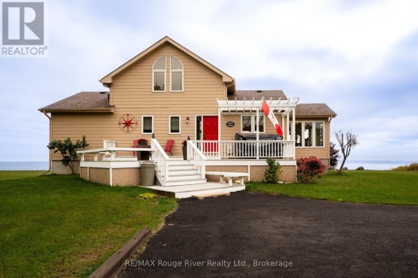 View of the tan house exterior from the driveway, showing the white deck, red door, and treed surroundings. - 455 Lakeshore Road, Alnwick/haldimand