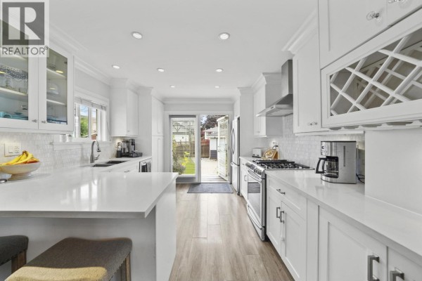 Modern white kitchen with stainless steel appliances, marble backsplash, and view into dining and living areas. - 4560 Pender Street, Burnaby