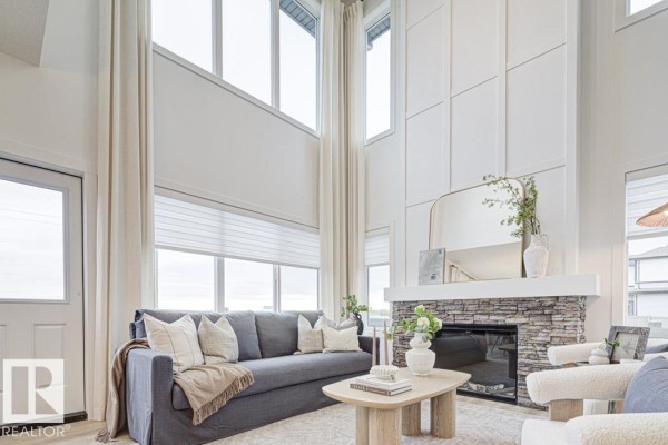Living room with high ceilings, a stone fireplace, a gray sofa, and large windows. - 4570 Chegwin Wd Sw, Edmonton