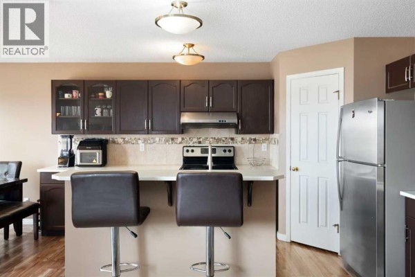 Kitchen with dark brown cabinets, stainless steel refrigerator, island with stools, and a closed white door. - 459 Citadel Meadow Bay Nw, Calgary