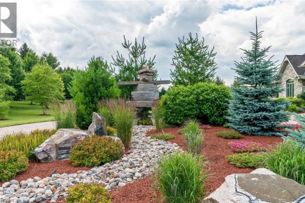 Decorative stone and rock garden with an inukshuk sculpture, mature trees, and manicured shrubs. - 46 Anita Drive, St  Clements