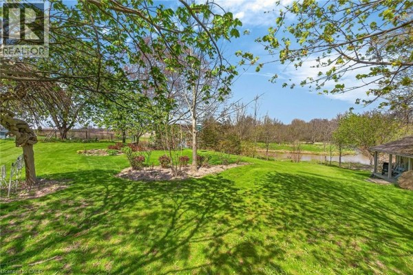 Backyard view of a large green lawn with mature trees, a small pond, and a wooden gazebo. - 46 Erie Street N, Selkirk