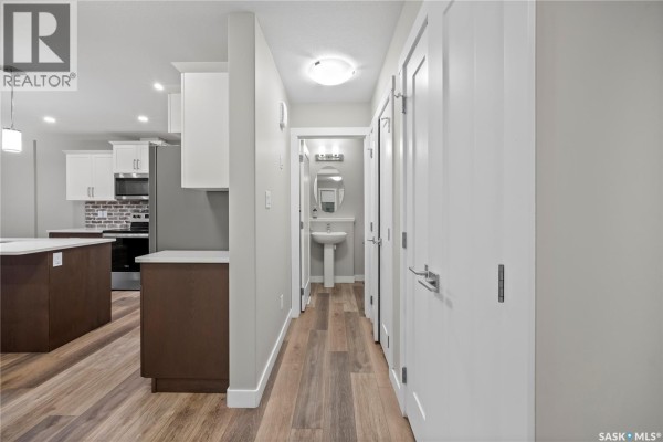 Hallway view showing doors, a pedestal sink, and a partial kitchen with white cabinets. - 4608 Mutrie Crescent, Regina