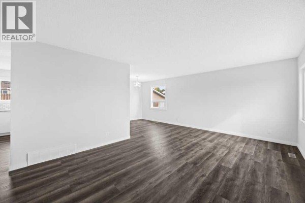 Empty living room with dark wood-look flooring and white walls. - 4628 Marcombe Road Ne, Calgary