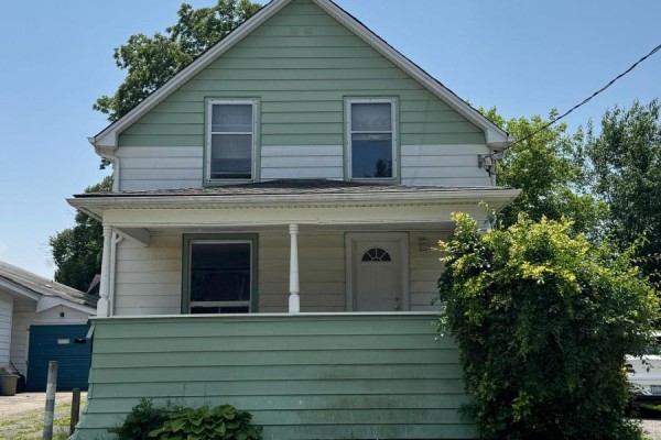 Front facade of a two-story house with white and green siding, a covered porch, and lush green shrubbery. - 4680 Ryerson Crescent, Niagara Falls