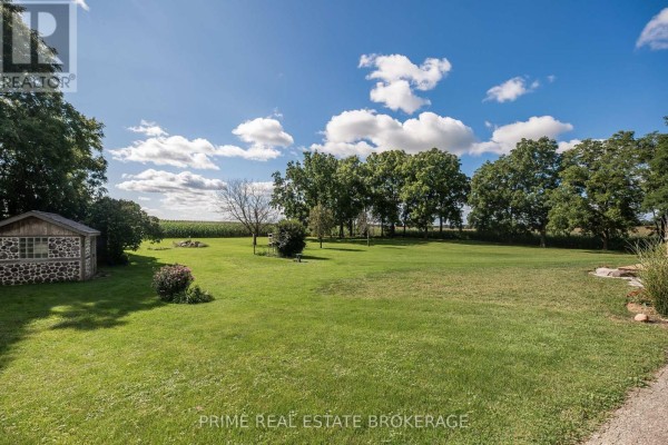 Spacious green lawn with mature trees, a stone shed, and distant fields. - 46821 Mapleton Line, Aylmer