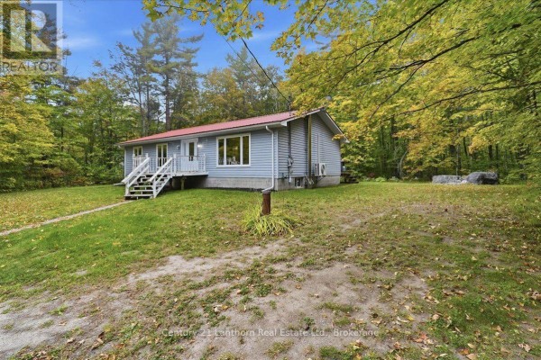 Side view of a light blue house with a red metal roof, set in a wooded area with fall foliage. - 47 Reynolds Road, Madoc