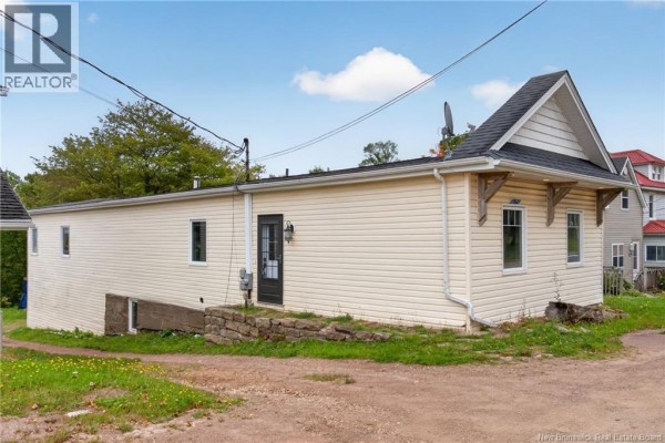 A beige residential building with a peaked roof, showing a side door, windows, and rough stone foundation. - 471 & 475 Centrale, Memramcook