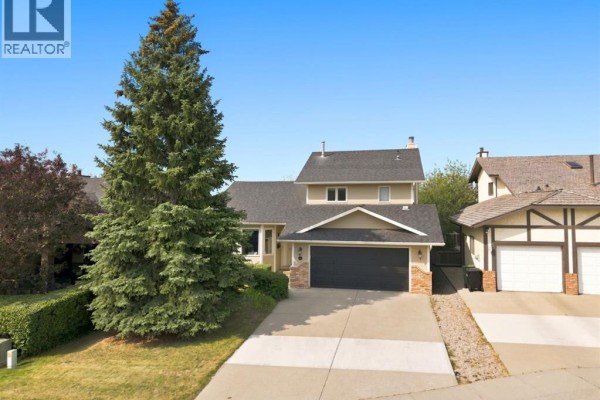 Side view of a beige house with a large evergreen tree in the front yard. - 473 Hawkford Way Nw, Calgary