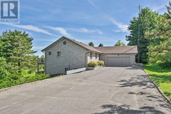 Grey brick house with a double car garage and a paved driveway surrounded by green trees under a blue sky. - 473 Sunset Crescent, Alnwick/haldimand