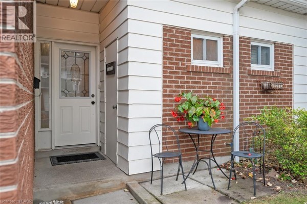 Front entrance of a townhouse with a white door, a small table and chairs, and brick and siding exterior. - 473 Woodview Road Unit# 2, Burlington