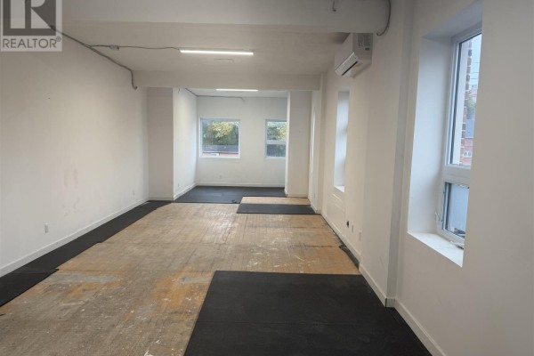 Empty commercial space with white walls, exposed wooden floor, and windows. Black mats cover some floor sections. - 478 Queen St, Sault Ste  Marie