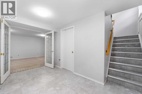Finished basement area with grey flooring, white walls, a staircase, and double glass doors. - 479 Bracewood Crescent Sw, Calgary