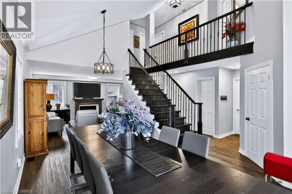 Open-concept dining and living area with a large dining table, fireplace, and dark wood railing on staircase. - 48 Holkham Avenue, Ancaster