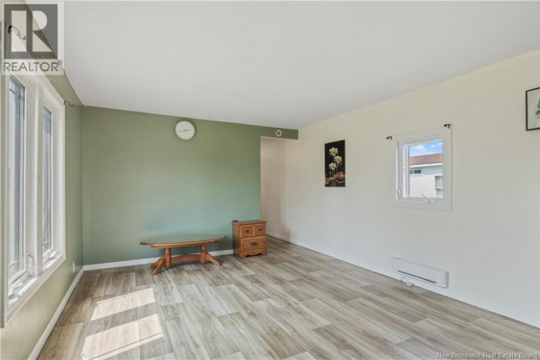Living room with light green and white walls, two windows, light wood laminate flooring, a small wooden bench, and a nightstand. - 48 Rue Des Copains, Richibucto