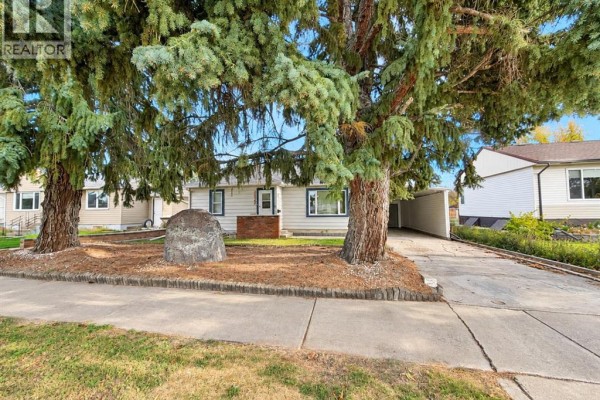 View of the house from the sidewalk, showing the front yard with two large trees. - 4825 51 Avenue, Taber