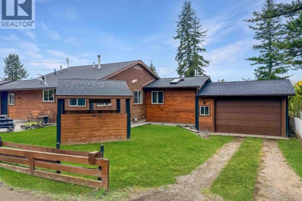 Side view of the wooden house showing a driveway, grass, and fence. - 4832 Clarke Avenue, Lac La Hache