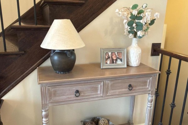 Close-up of a wooden console table with drawers, a lamp, a framed photo, and a vase. - 4871 Northgate Crescent, Lincoln