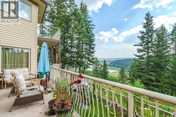 Balcony view from a beige house overlooking a valley with a lake and scattered trees under a cloudy sky. - 4882 Gloinnzun Crescent, 108 Mile Ranch