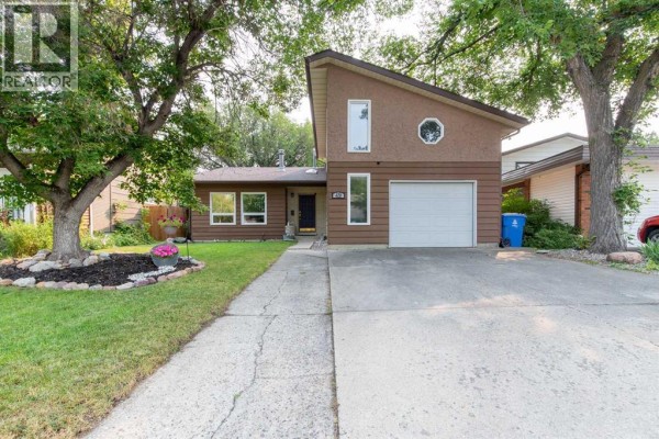 Exterior front view of a two-storey brown house with an attached garage, surrounded by trees. - 49 Iroquois Crescent W, Lethbridge