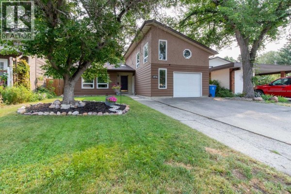View of the house exterior from the driveway, showing trees, a garage, and part of the yard. - 49 Iroquois Crescent W, Lethbridge