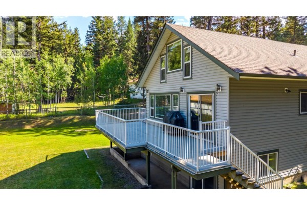 A large grey deck with white railings wraps around the back of a grey sided house with large windows. - 4931 Kyllo Road, 108 Mile Ranch