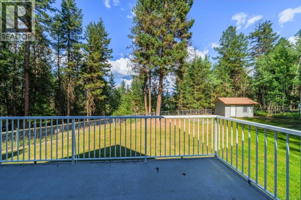 View from a deck of a grassy backyard surrounded by tall pine trees with a small shed in the distance. - 4931 Kyllo Road, 108 Mile Ranch
