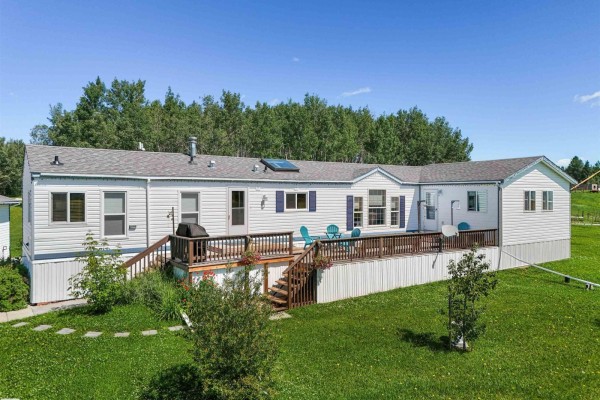 Rear exterior view of a white mobile home with a large wooden deck featuring blue chairs and a grill. - 49327 Rge Rd 53, Rural Brazeau County