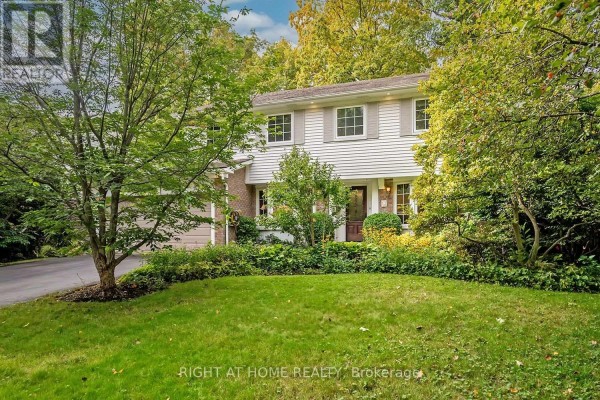 Side view of a two-story home with white siding and brick accents, surrounded by mature trees and lush landscaping. - 494 Dorland Road, Oakville