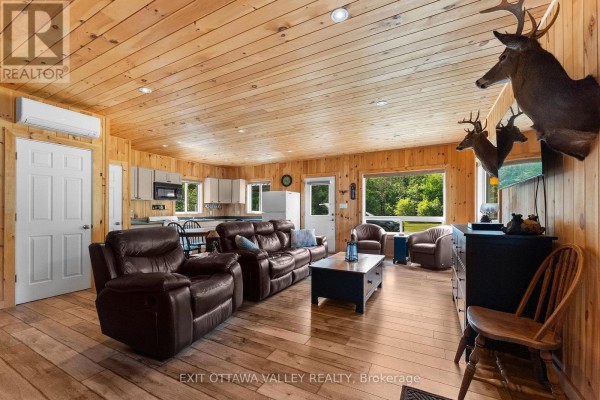 Living room with wood-paneled walls, plush seating, kitchen area, and views of trees. - 495 Gravel Pit Lane, Wollaston