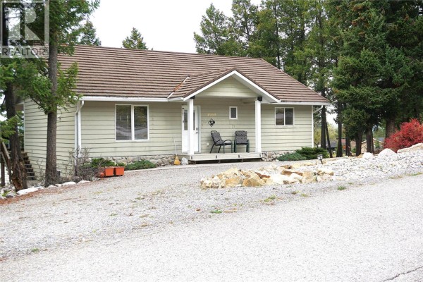 Slightly angled front view of a light green, single-story house with a brown roof and trees. - 4969 Mountain Side Road, Fairmont Hot Springs